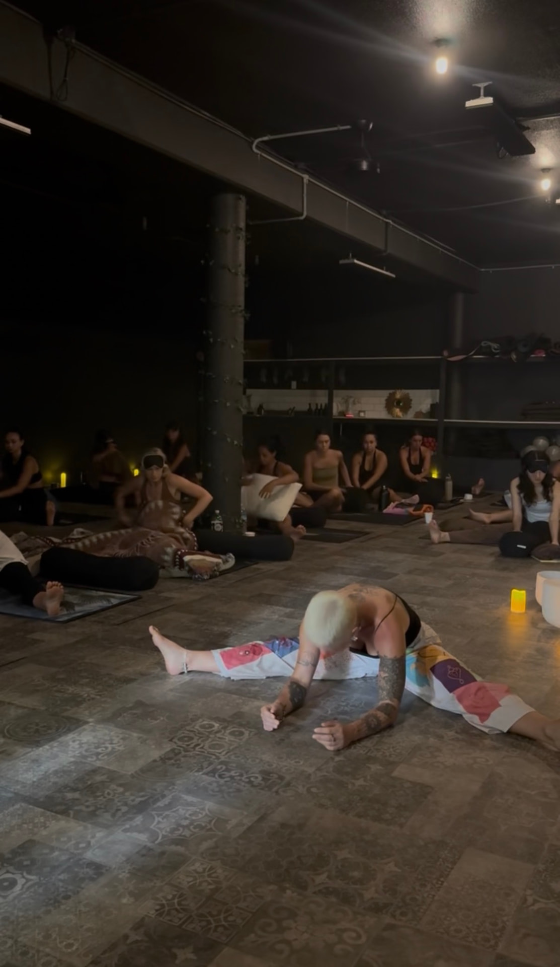 A candle-lit studio session with Sharleen in a wide forward fold on the floor, group seated around her