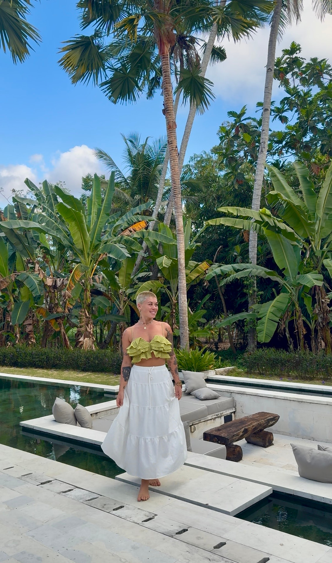 Sharleen standing barefoot in a tropical garden beside a pool, palms and banana plants behind her