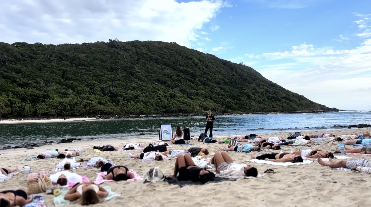 Sharleen facilitating a large beach session with a group lying on the sand below a green headland